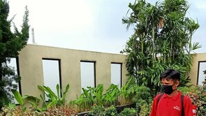 A man in a red shirt standing in a patio area with wicker chairs, a glass table, and various plants. - Grand Kasira (Jakarta)