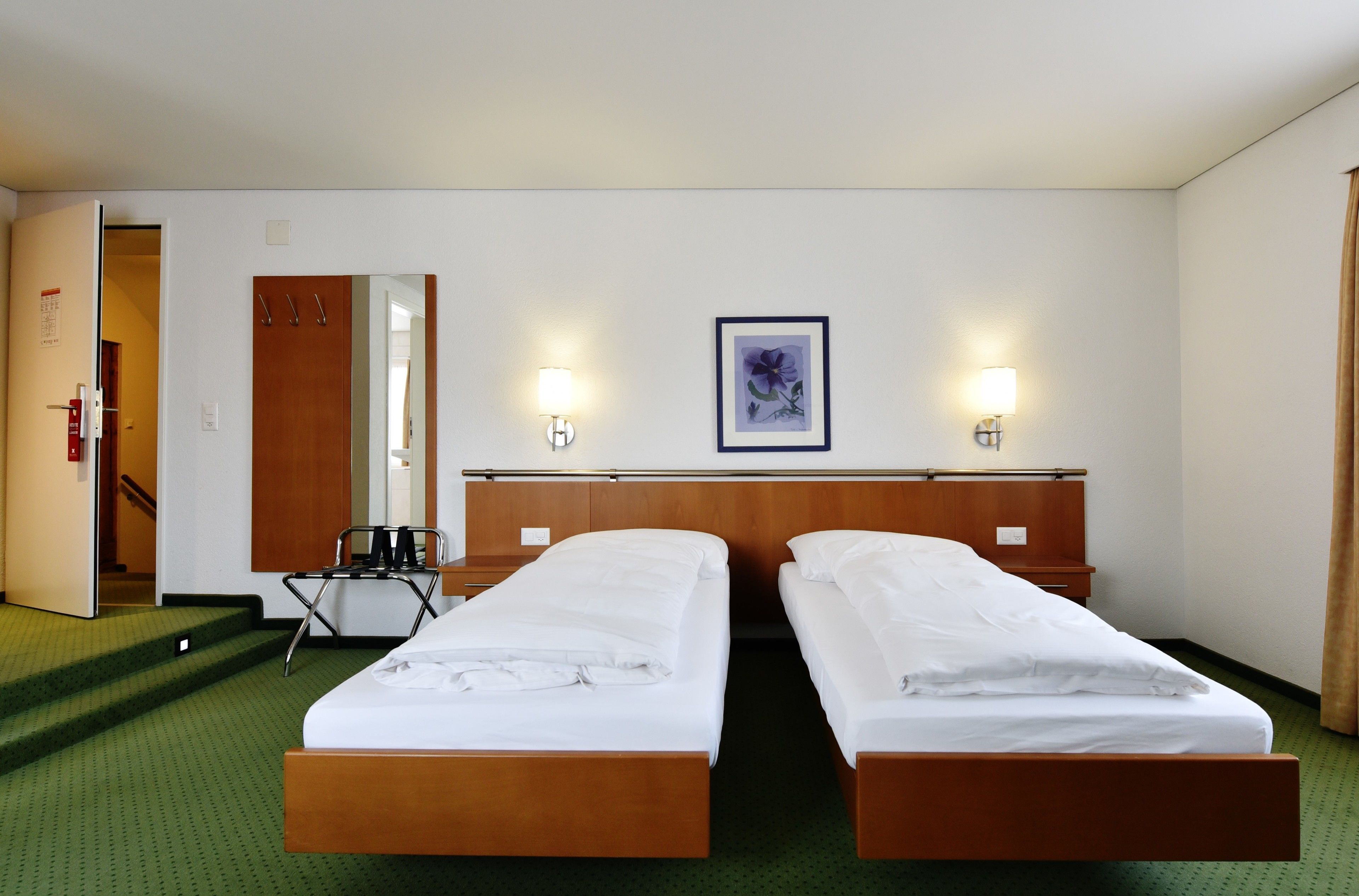 Habitación de hotel con dos camas individuales, cabecera de madera, un escritorio pequeño con una silla, un espejo y dos lámparas fijadas a la pared.