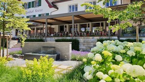 A hotel with a covered outdoor seating area, a stone water feature, and lush greenery.