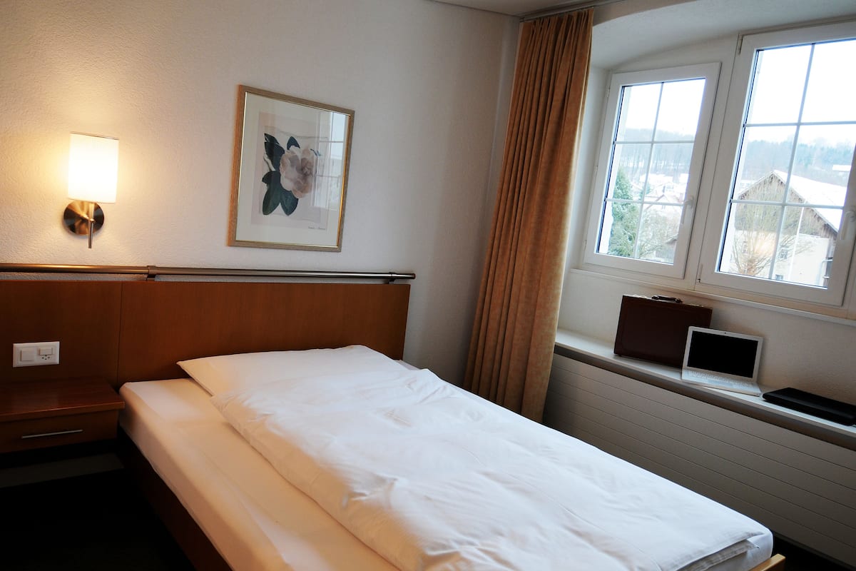 Een hotelkamer met een opgemaakt bed, een bedlampje, een ingelijst schilderij aan de muur, een raam met gordijnen en een laptop op het raamkozijn.