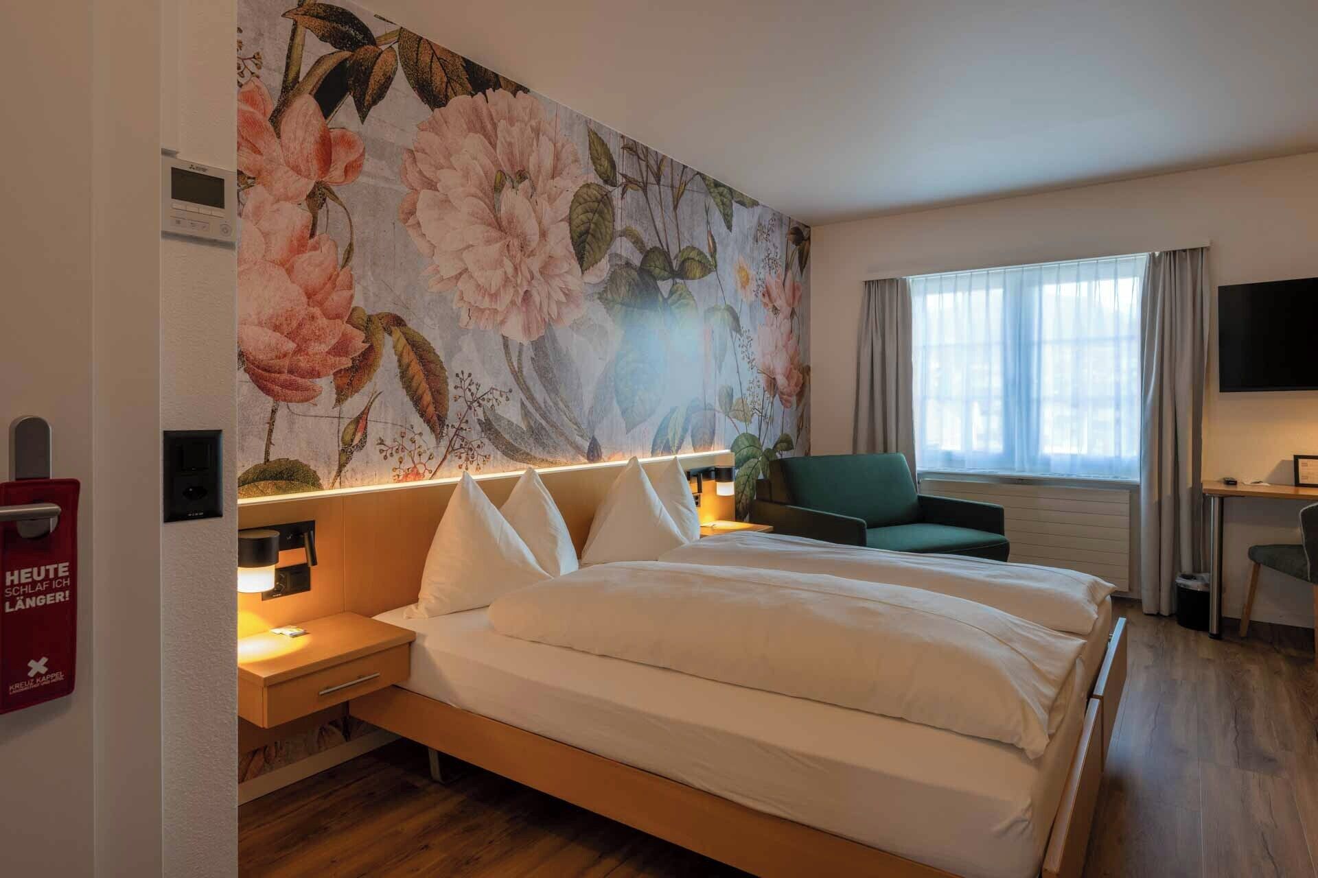A hotel room with a large floral mural, a bed with white linens, a green armchair, and a desk with a television.