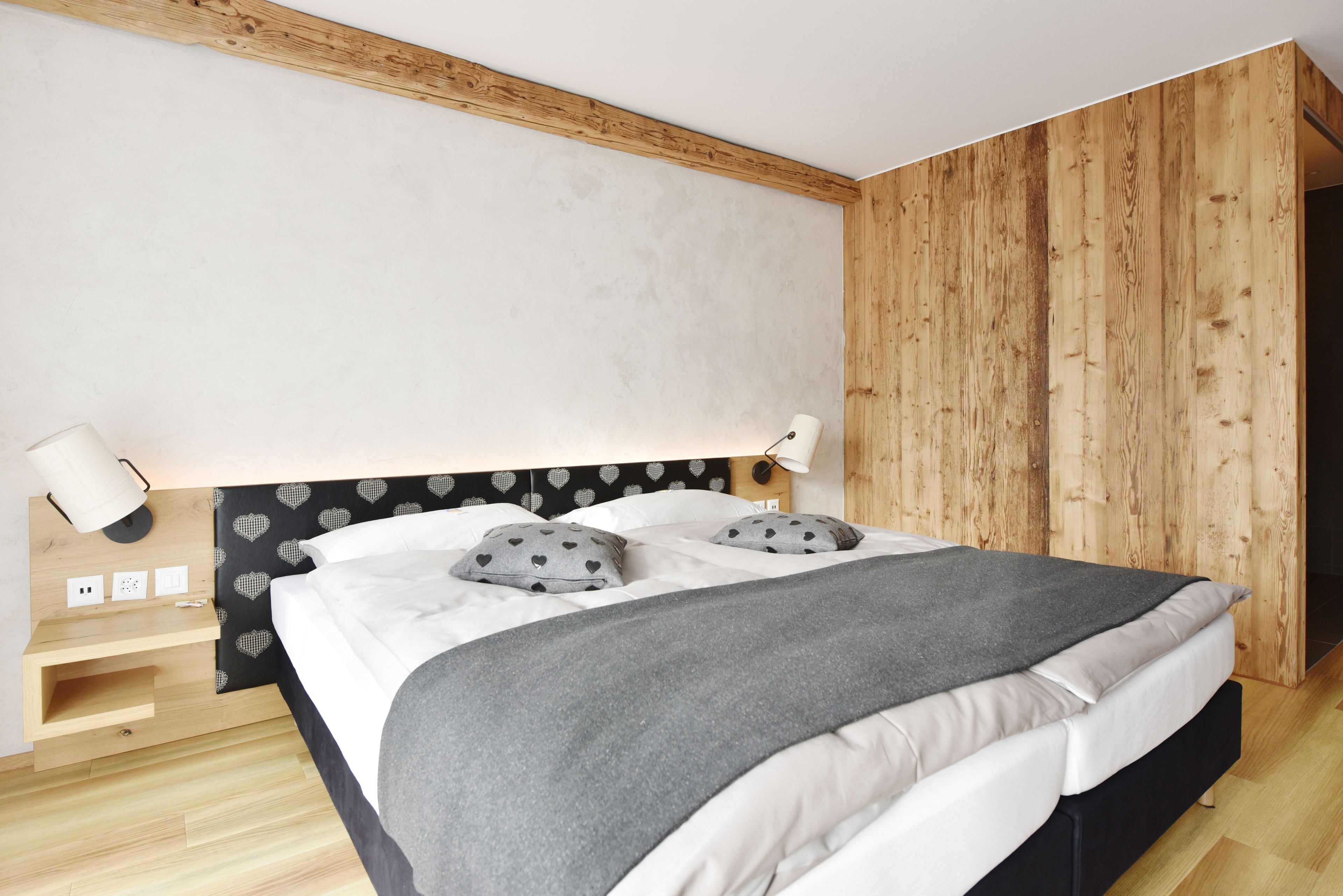 A modern bedroom with a wooden headboard, a bedside table with a lamp, and a bed with a grey blanket and heart-patterned pillows.