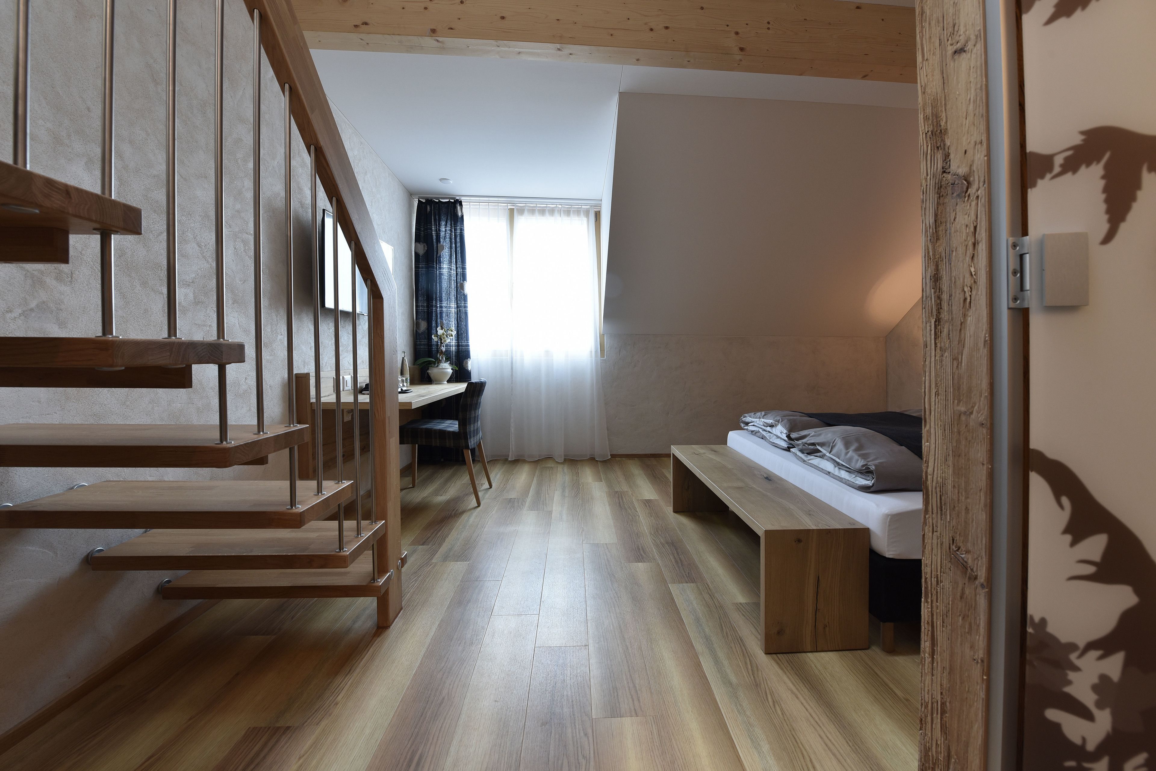 A modern room with a wooden staircase, a desk with a chair, a bed, and a window with curtains.