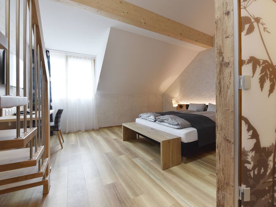 A modern bedroom with a sloped ceiling, a wooden staircase, a bed with a bench, and a desk.