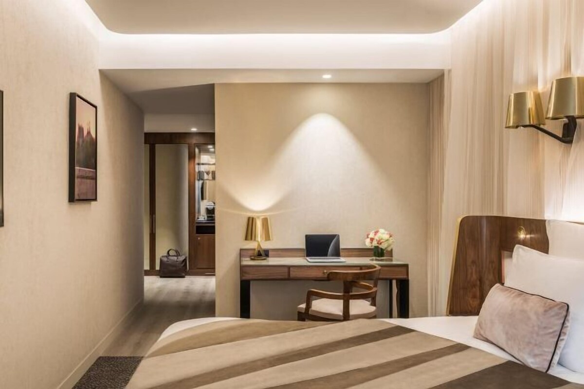 A hotel room with a bed, desk, chair, and a view of the hallway.