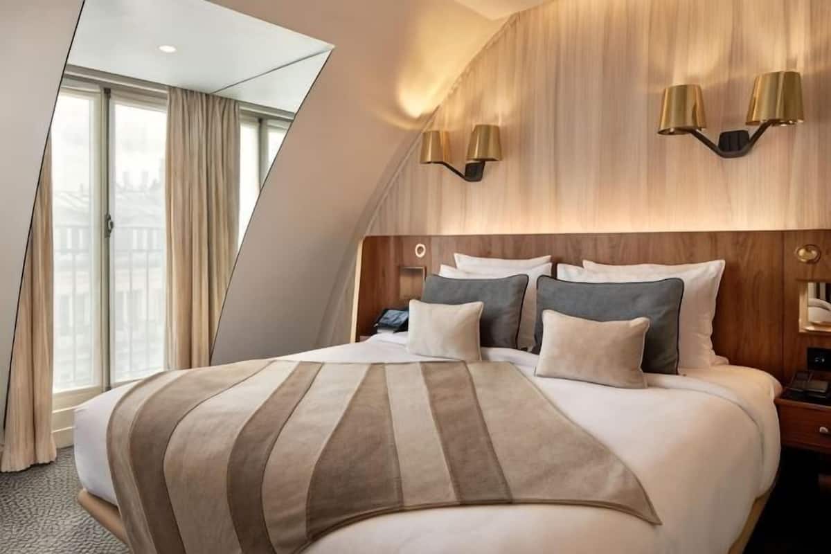 A hotel room with a large bed, a wooden headboard, two wall-mounted lamps, and a view of the city through the window.