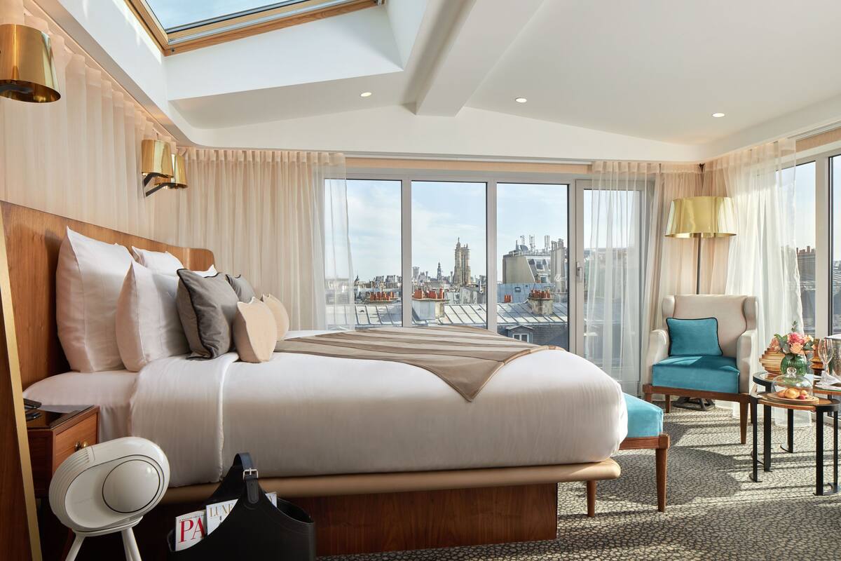 A modern hotel room with a large bed, a sitting area with a chair and table, and a city view through large windows.