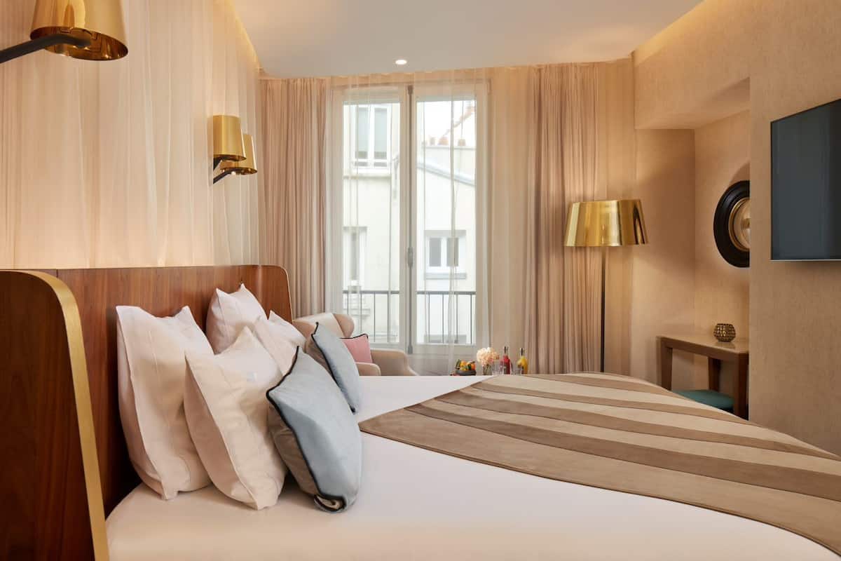 A hotel room with a large bed, a bedside table, a lamp, and a view of buildings through the window.