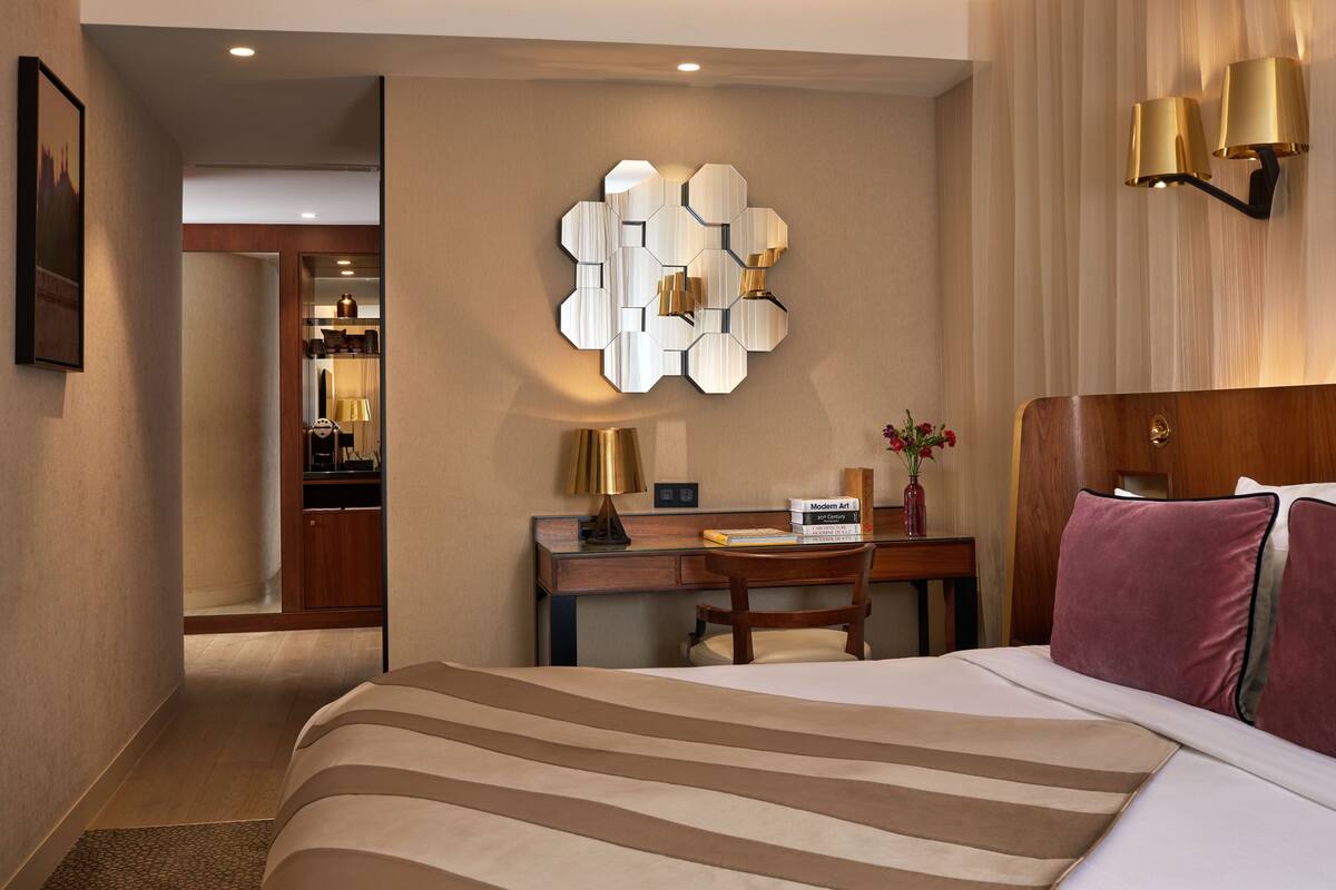 A hotel room with a large bed, a desk with a chair, a wall-mounted lamp, and a decorative wall piece.