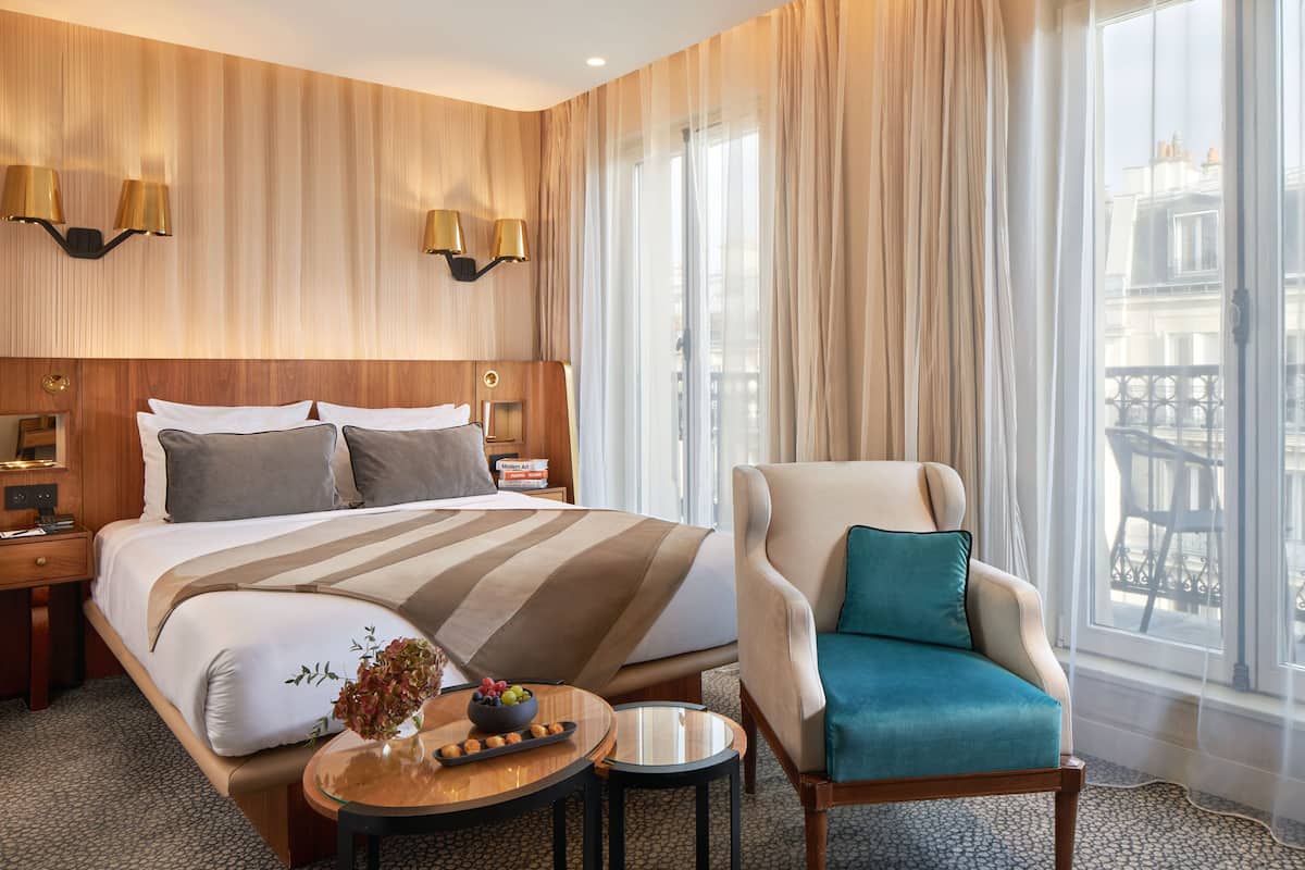 A hotel room with a large bed, a sitting area with a chair, a small table with a vase and fruit, and a view of the city through large windows.