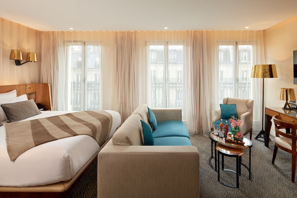A hotel room with a bed, a sofa, a chair, a small table with drinks and flowers, and large windows with curtains.