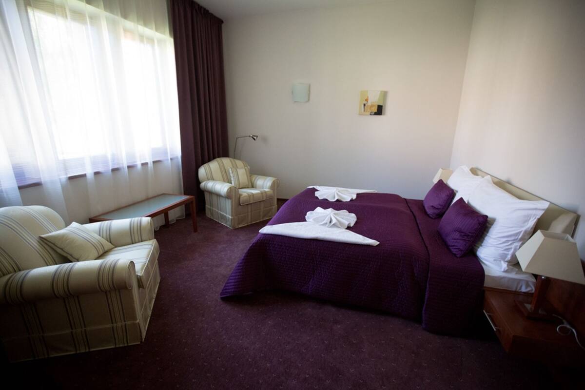 Een hotelkamer met een groot bed, twee fauteuils, een tafeltje en een raam met gordijnen.