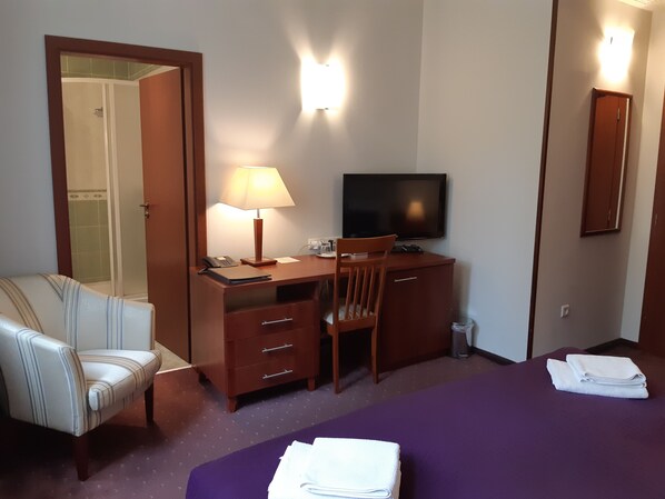 A hotel room with a bed, a desk with a chair, a television, a lamp, and a bathroom with a shower.