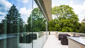 Een modern terras buiten, met glazen wanden, tuinmeubilair en uitzicht op bomen en de lucht.