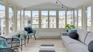 Une pièce lumineuse, baignée de soleil, avec de grandes fenêtres, un canapé gris, un fauteuil turquoise, une chaise en osier noire et une petite table sur laquelle est posée une plante.