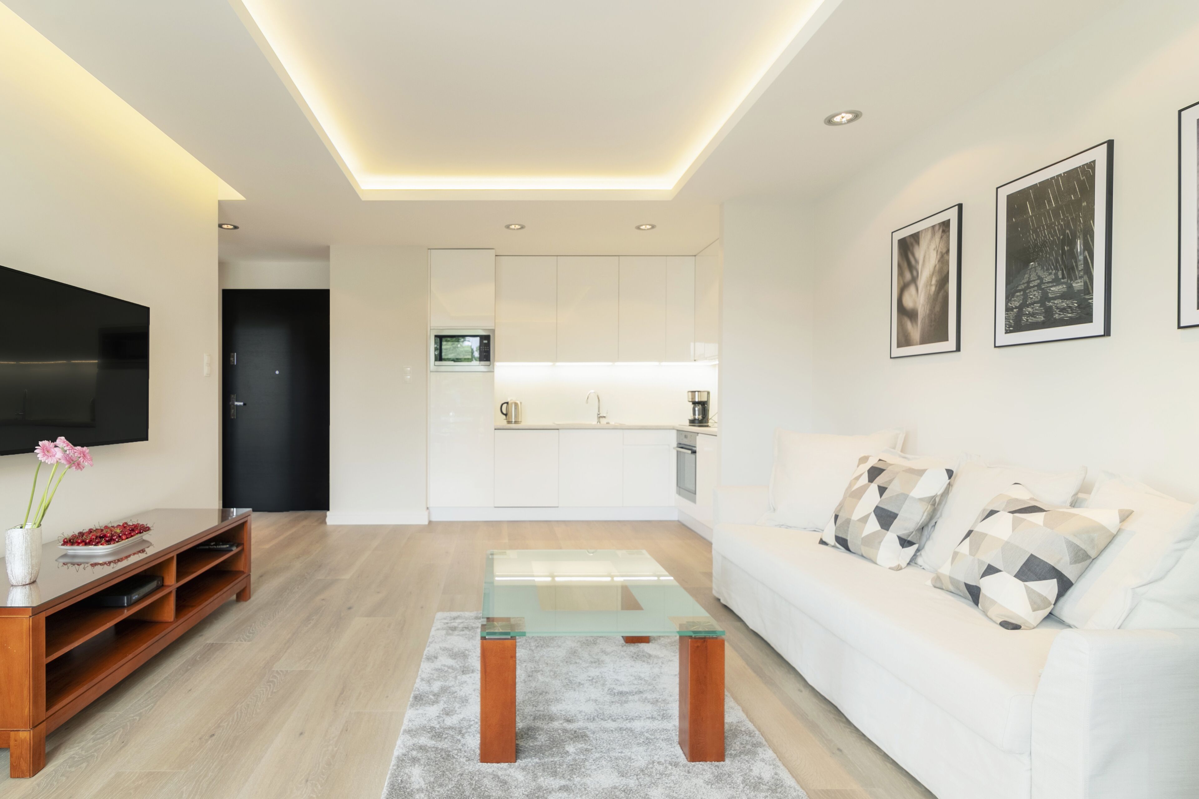A modern living room with a glass coffee table, white sofa, and a flat-screen TV.