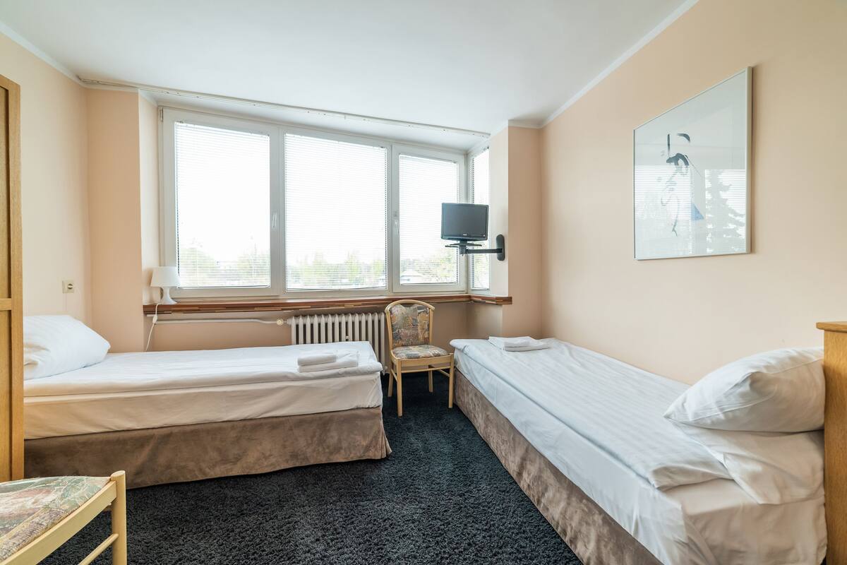 A room with two beds, a chair, a radiator, a TV, and a window with blinds.