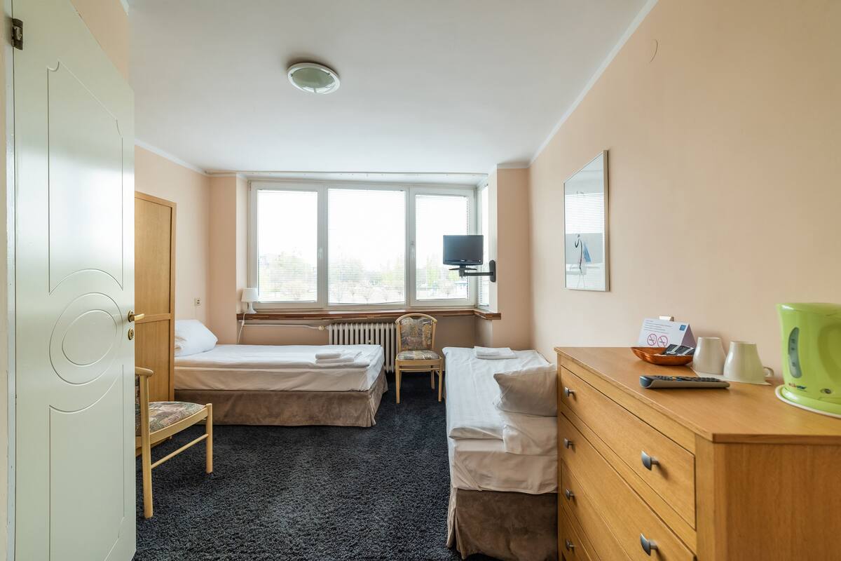A hotel room with two beds, a chair, a dresser, a radiator, a television, and a window with blinds.