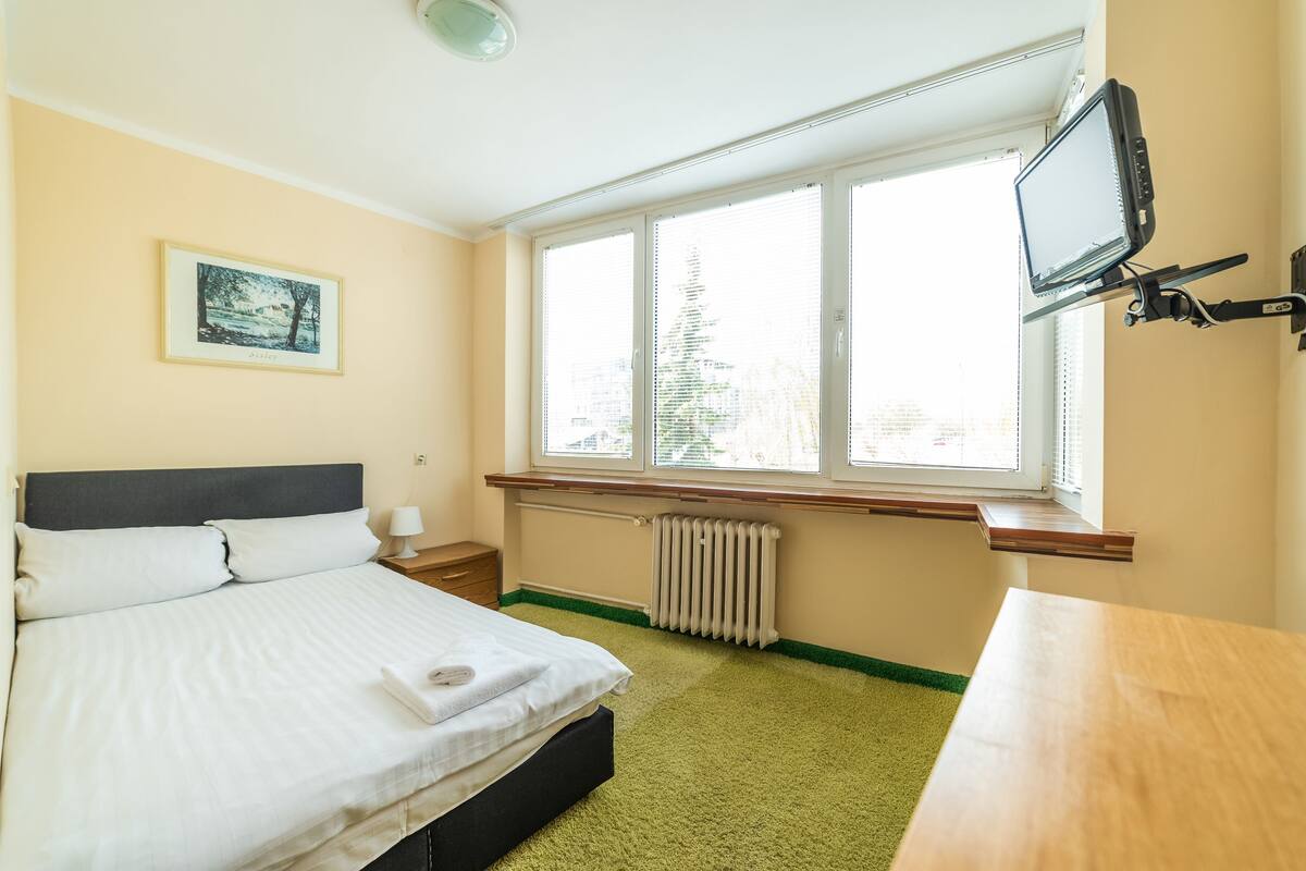 A bedroom with a bed, a window with blinds, a radiator, a bedside table with a lamp, and a TV mounted on the wall.