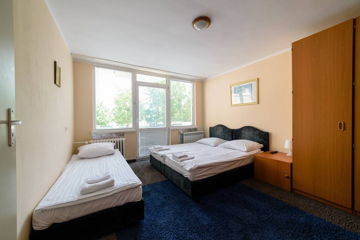 A room with two single beds, a wooden wardrobe, a nightstand with a lamp, and a window with blinds.
