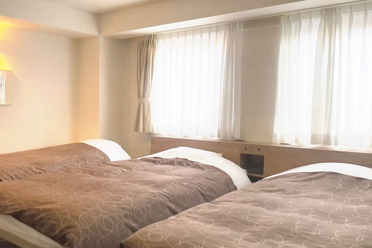 A hotel room with two beds, each with a patterned bedspread and pillows. There is a window with curtains, a bedside table, and a wall-mounted lamp.