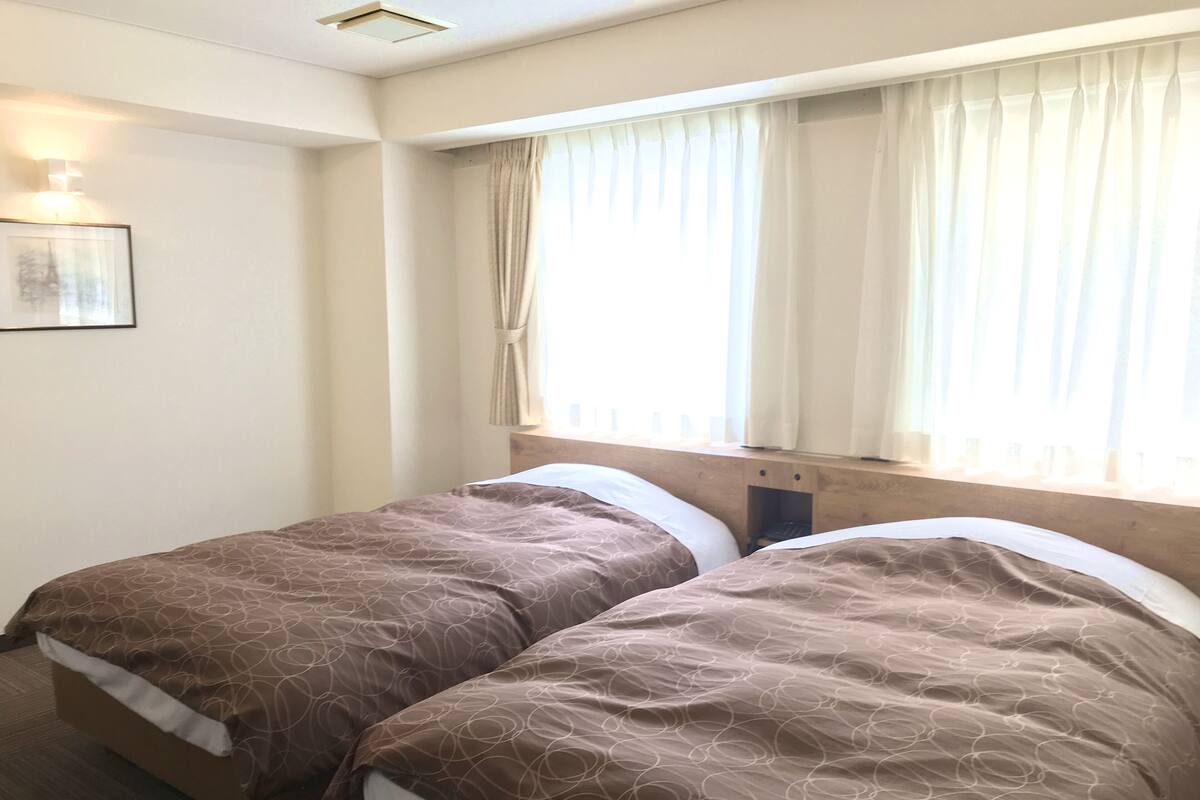 A hotel room with two beds, a window with curtains, a wall-mounted light, and a framed picture.