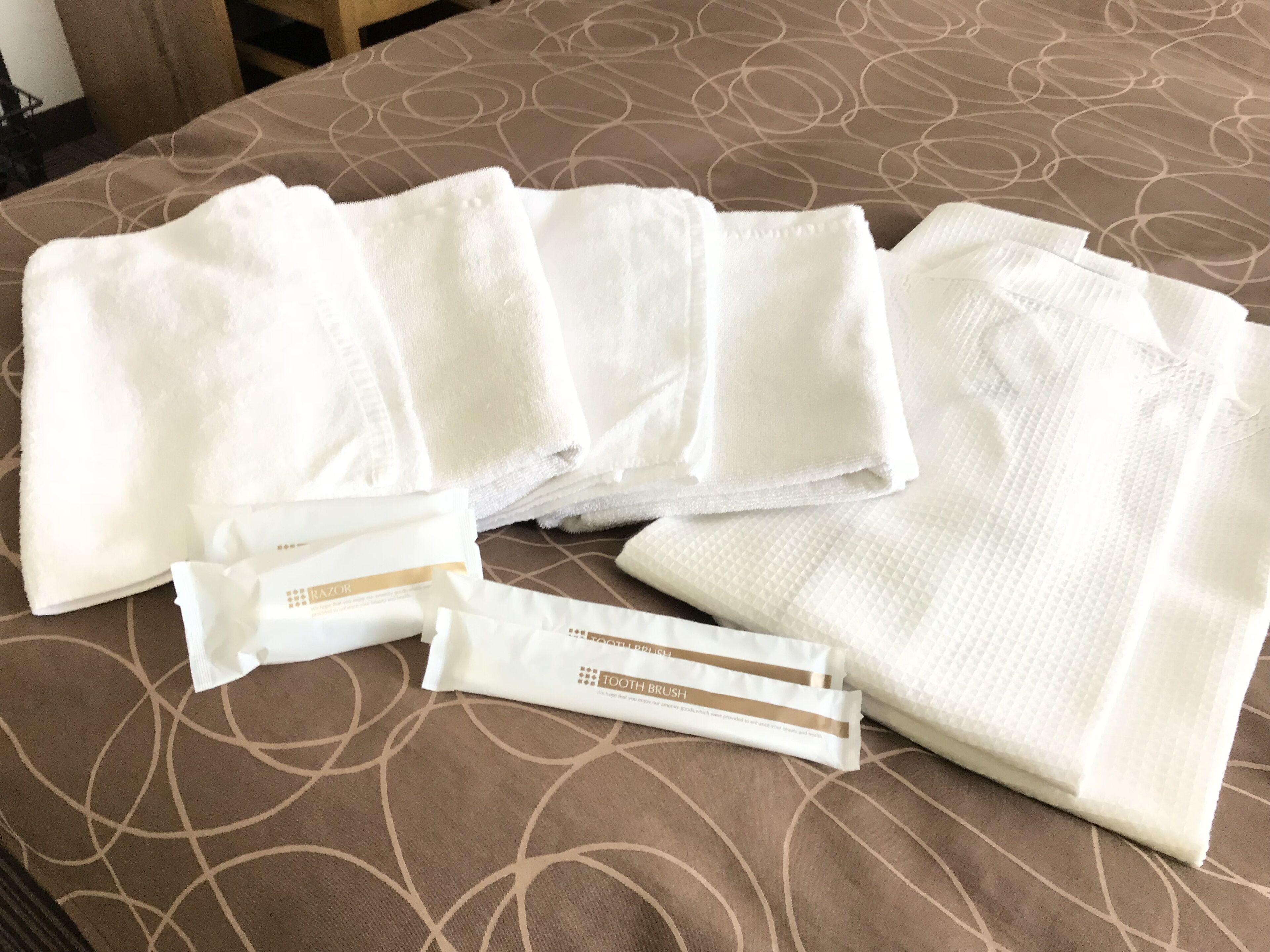 A bed with a brown patterned bedspread and several neatly folded white towels and a toothbrush.