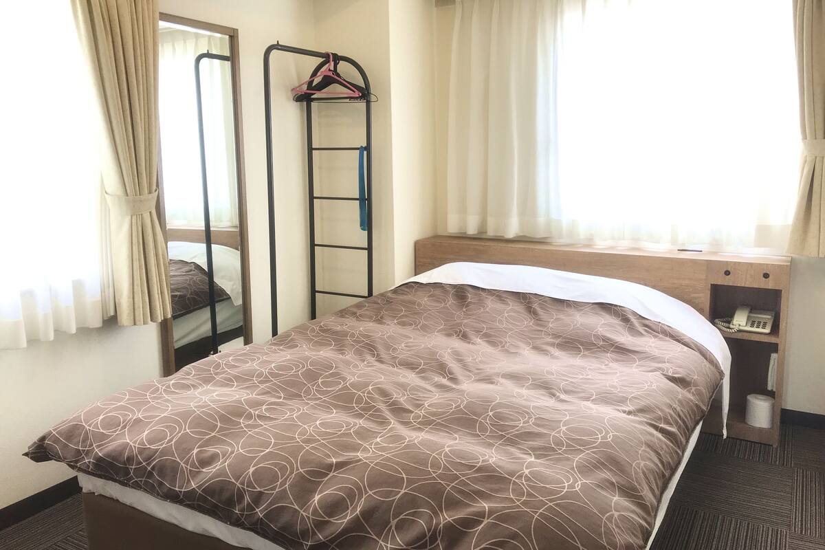 A single bed with a patterned bedspread, a bedside table with a telephone, a mirror, and a clothing rack.
