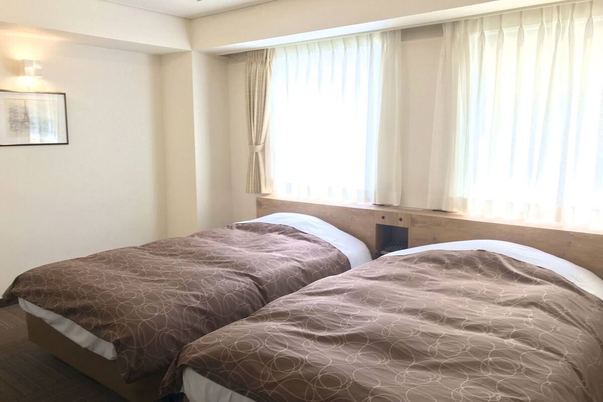 A hotel room with two beds, a window with curtains, a wall-mounted light, and a framed picture.
