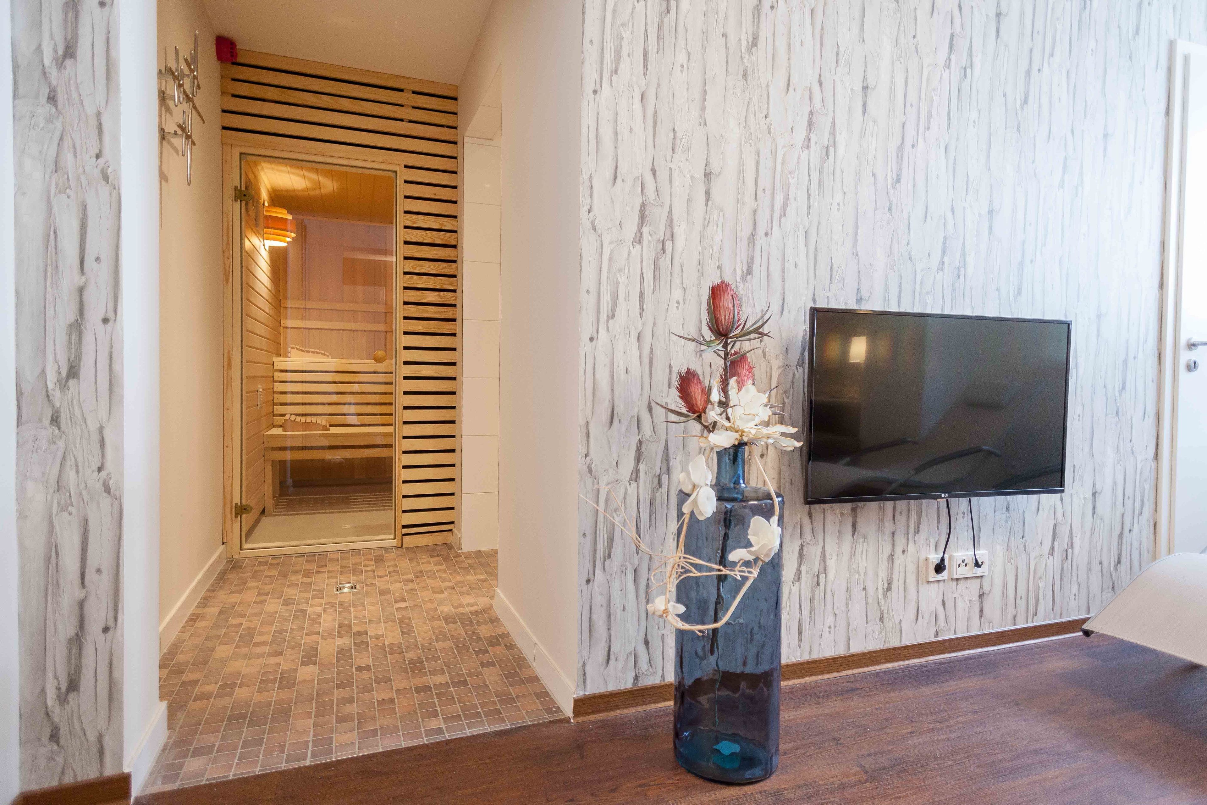 Ein modernes Interieur mit Holzboden, einer Glastür zum Duschbereich, einem an der Wand montierten Fernseher und einer Vase mit Blumen.