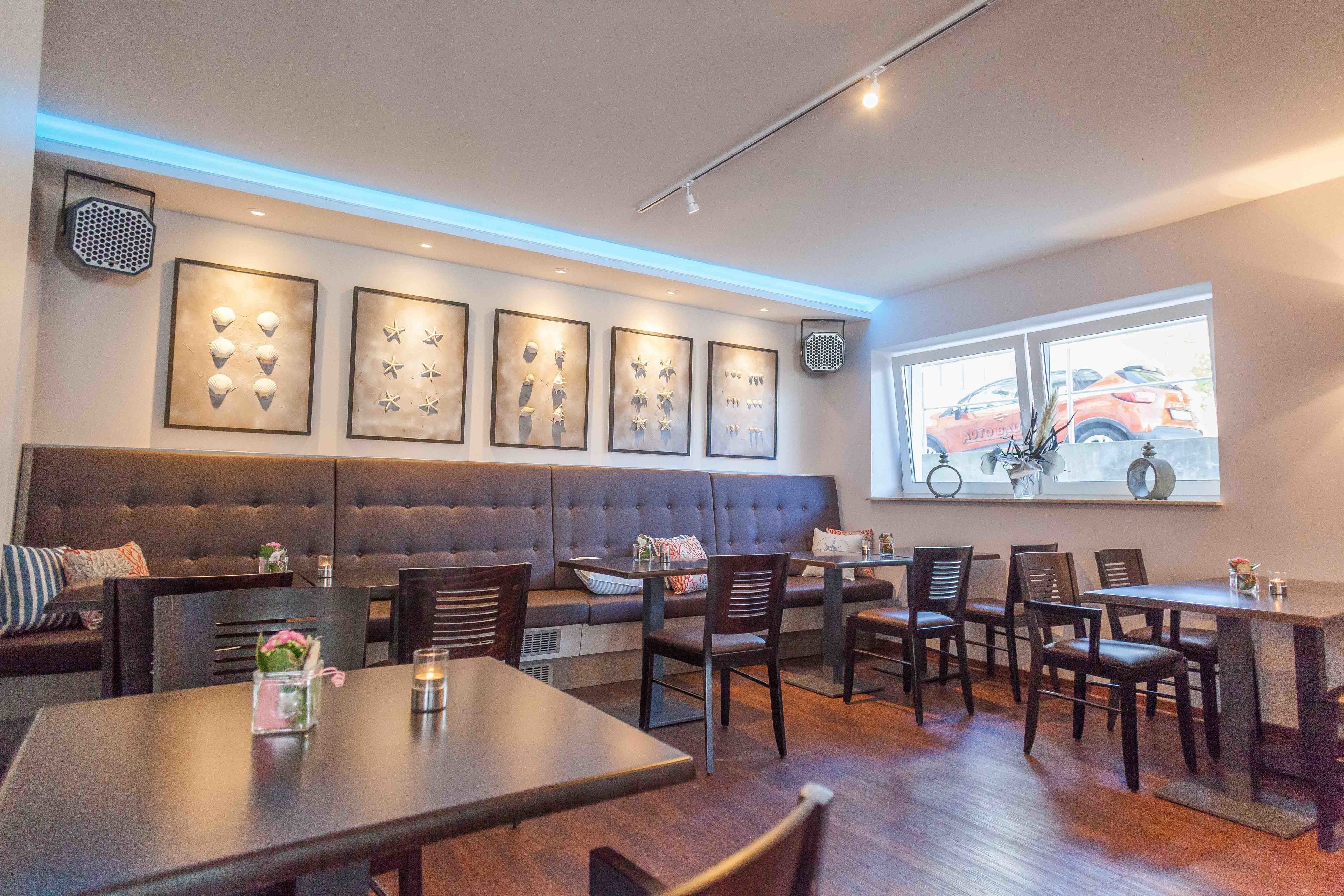 Un espace repas moderne avec des banquettes en cuir foncé, des tables et des chaises en bois. Des œuvres d’art encadrées ornent le mur, et une fenêtre offre une vue sur les voitures garées.