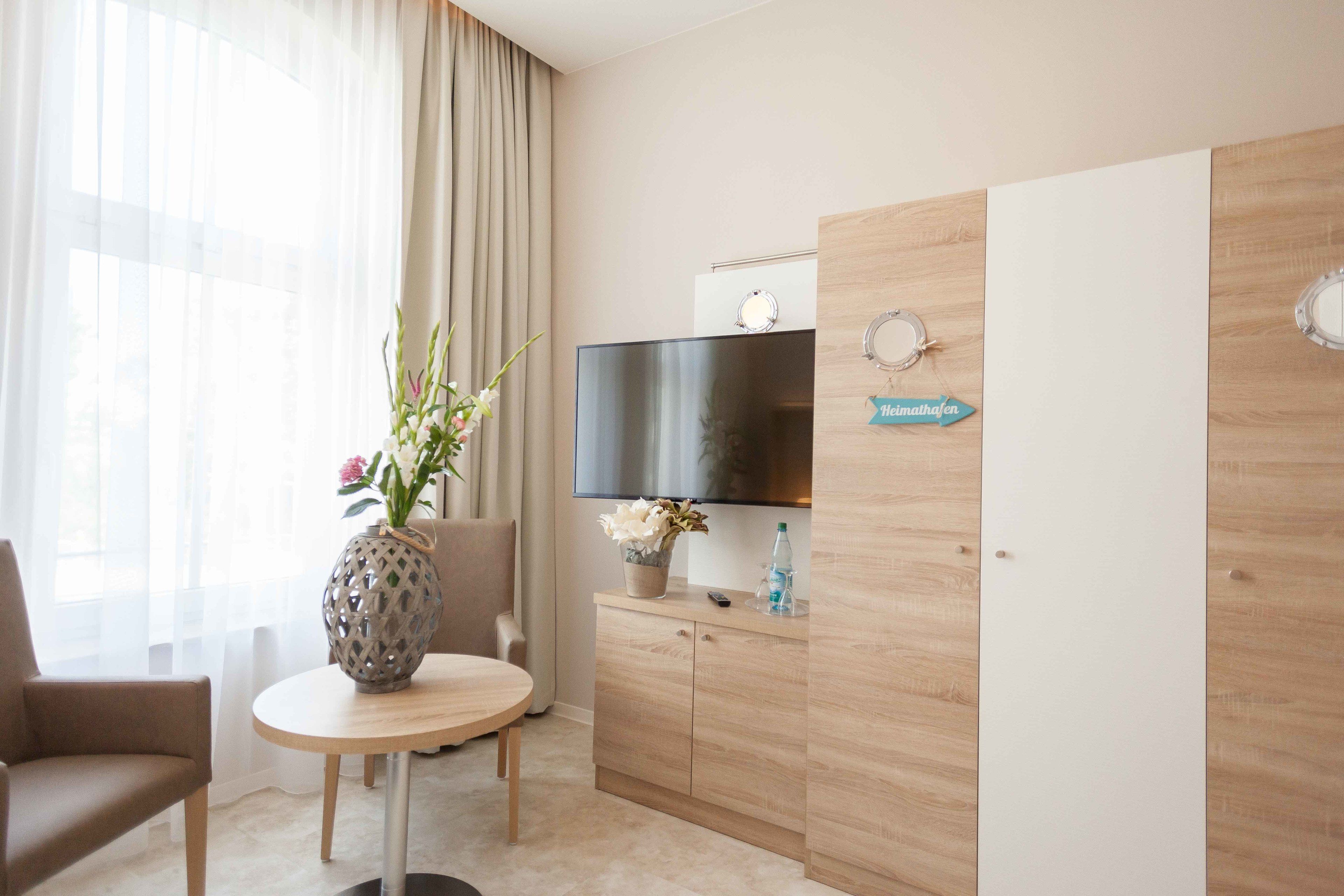 A hotel room with a round wooden table, a beige armchair, a TV on a wooden cabinet, and a vase with flowers on the table.