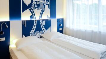 Habitación de hotel con una cama, pared decorada con diseño azul y blanco, una ventana con cortinas y una mesita de noche con lámpara.