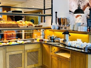 Un área de buffet con diferentes opciones de comida, una estación de café y un mostrador con vasos y platos.