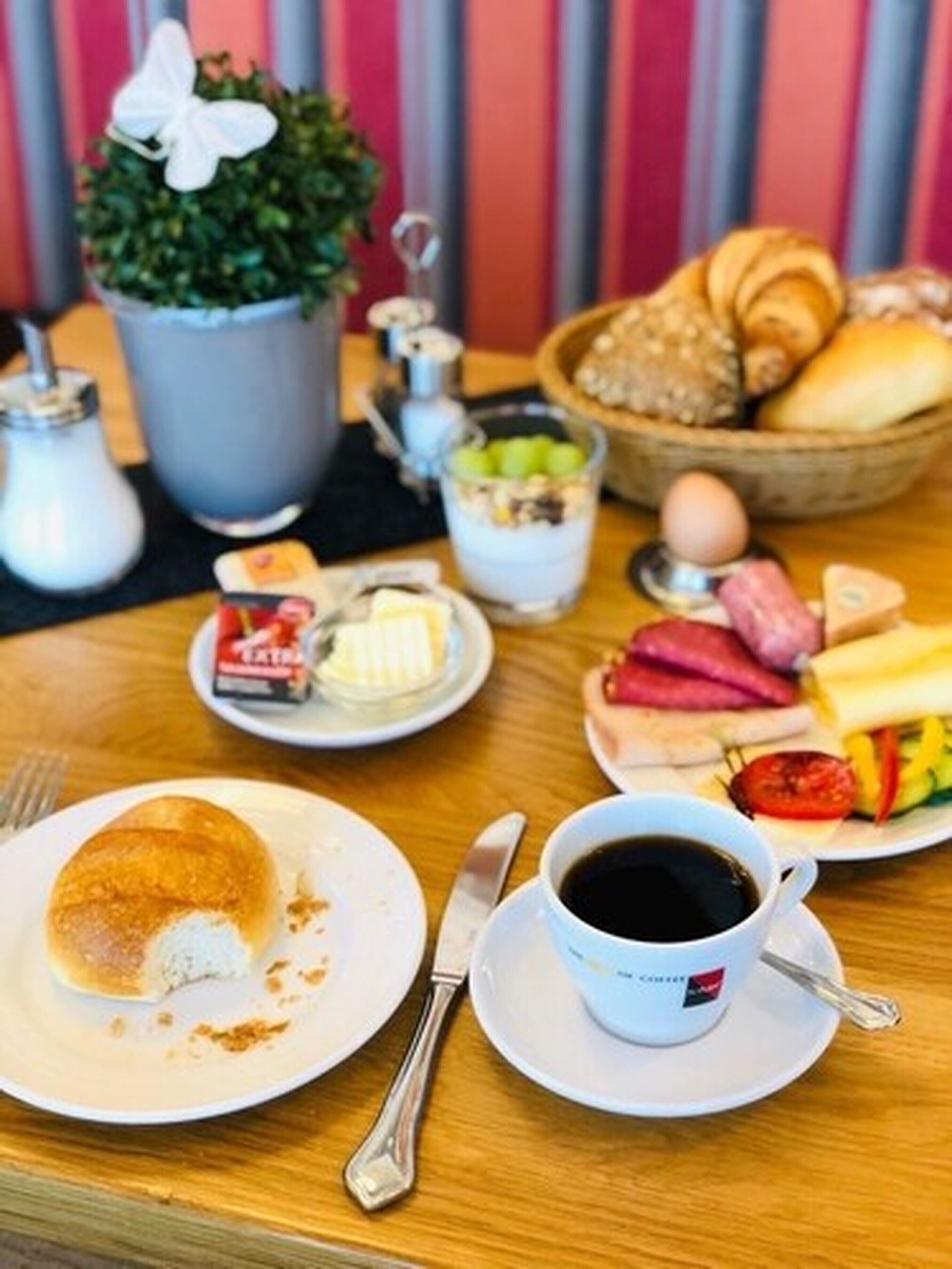 Taula de petit esmorzar amb un croissant, un plat de pa tallat, un bol de fruita, una tassa de cafè i un pot petit de mantega.