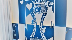 A hotel room with a large wall mural of a playing card, a bed with white bedding and blue pillows, a bedside table with a remote control, and a window with curtains. - Aparthotel Paderborn (Paderborn)