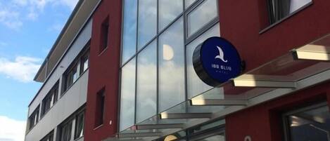 Egy modern épület, üveg homlokzattal, egy kék színű táblával, amelyen a „188 BLUE” felirat és egy madárlogó látható, valamint egy „2” szám, amely a bejáratot jelöli.