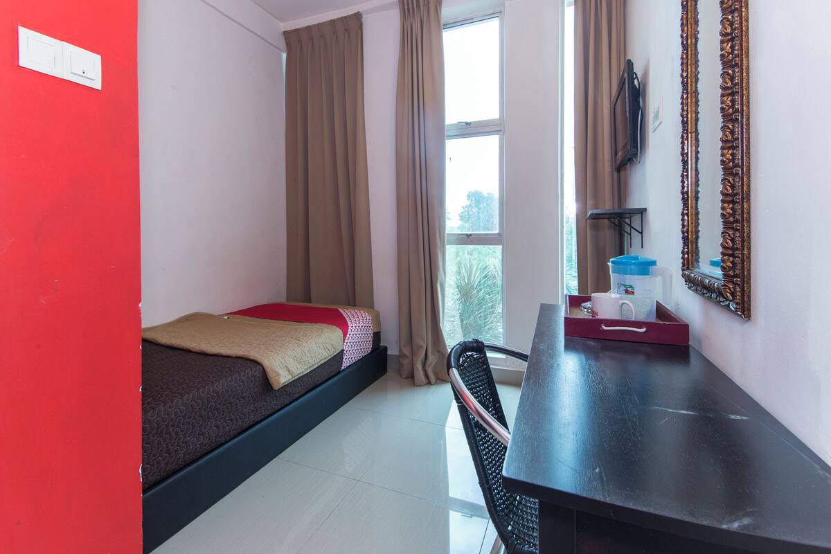 A small, single-bed room with a desk and chair, a window with curtains, and a mirror on the wall.