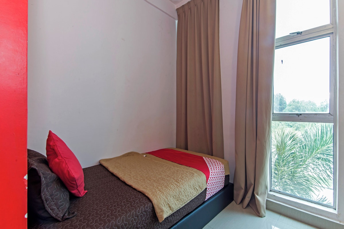 A small, single-bed room with a window, a red pillow, a beige blanket, and a patterned bedspread.