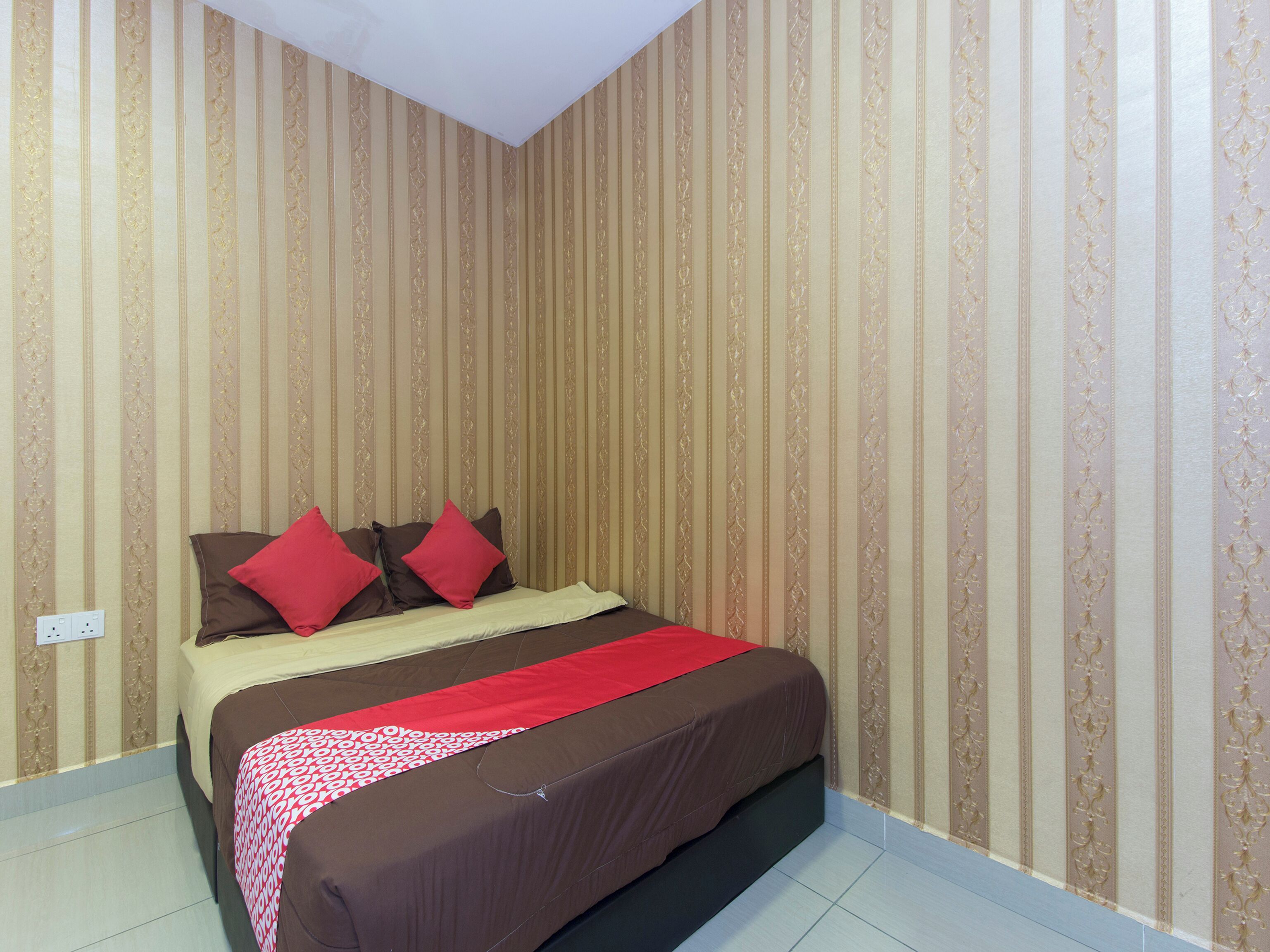 Een net opgemaakt bed met een bruinrood sprei, een rode deken en twee rode kussens. De kamer heeft gestreept behang en een wit plafond.