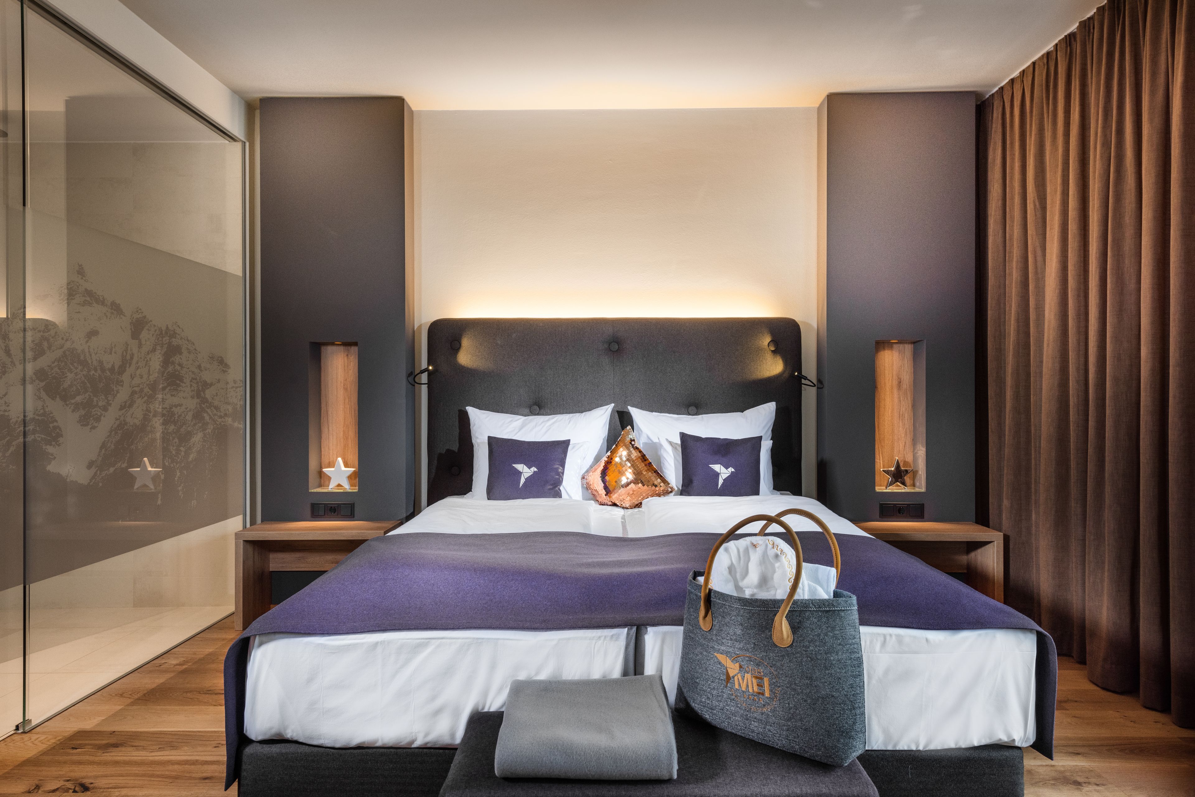 A modern hotel room with a large bed, a nightstand, a bedside table, a wall-mounted lamp, and a large window with a view of trees.