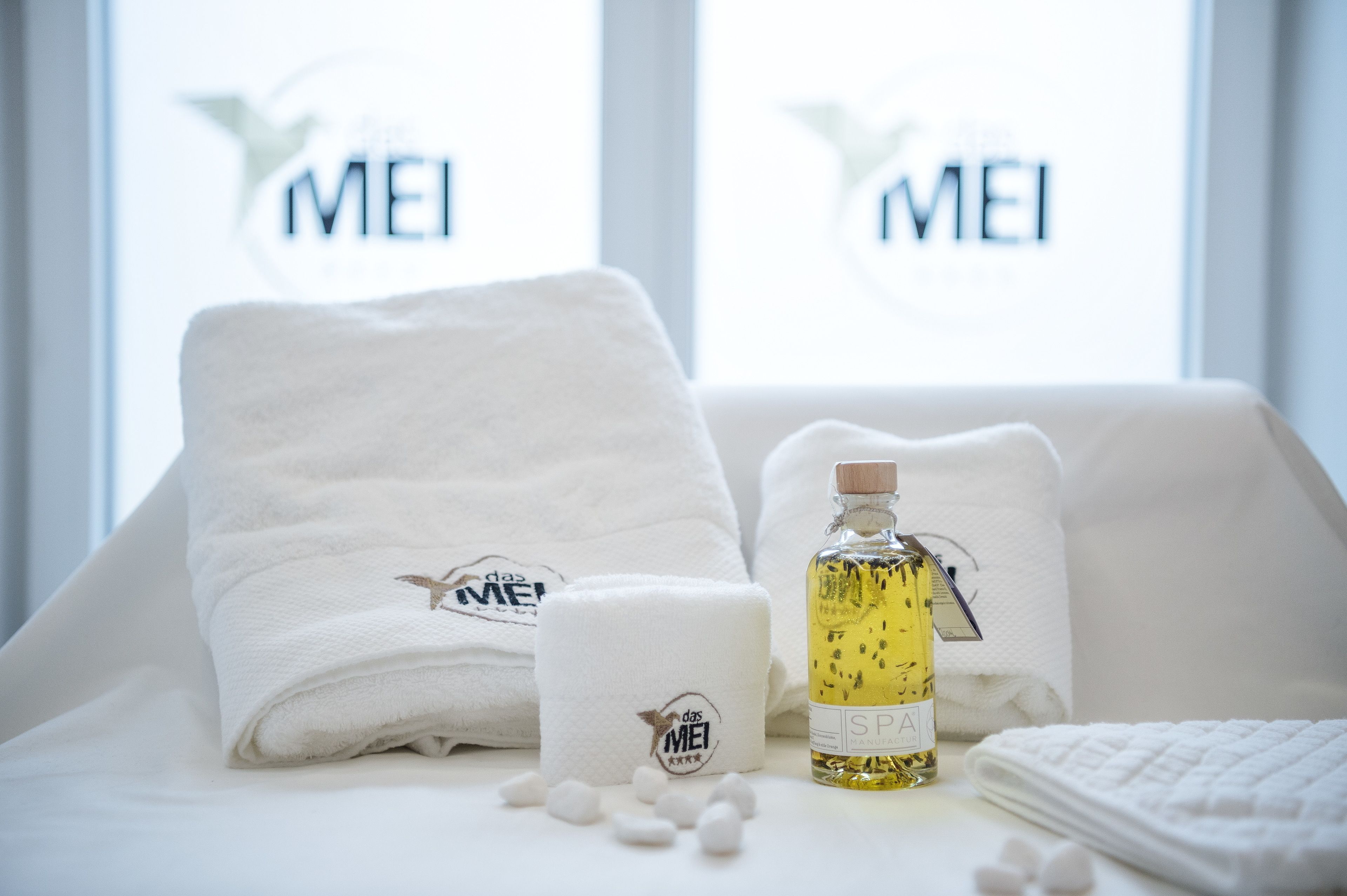 Uma casa de banho bem organizada com toalhas brancas, um frasco de óleo de SPA e uma pequena vela, tudo com a marca "das MEI".