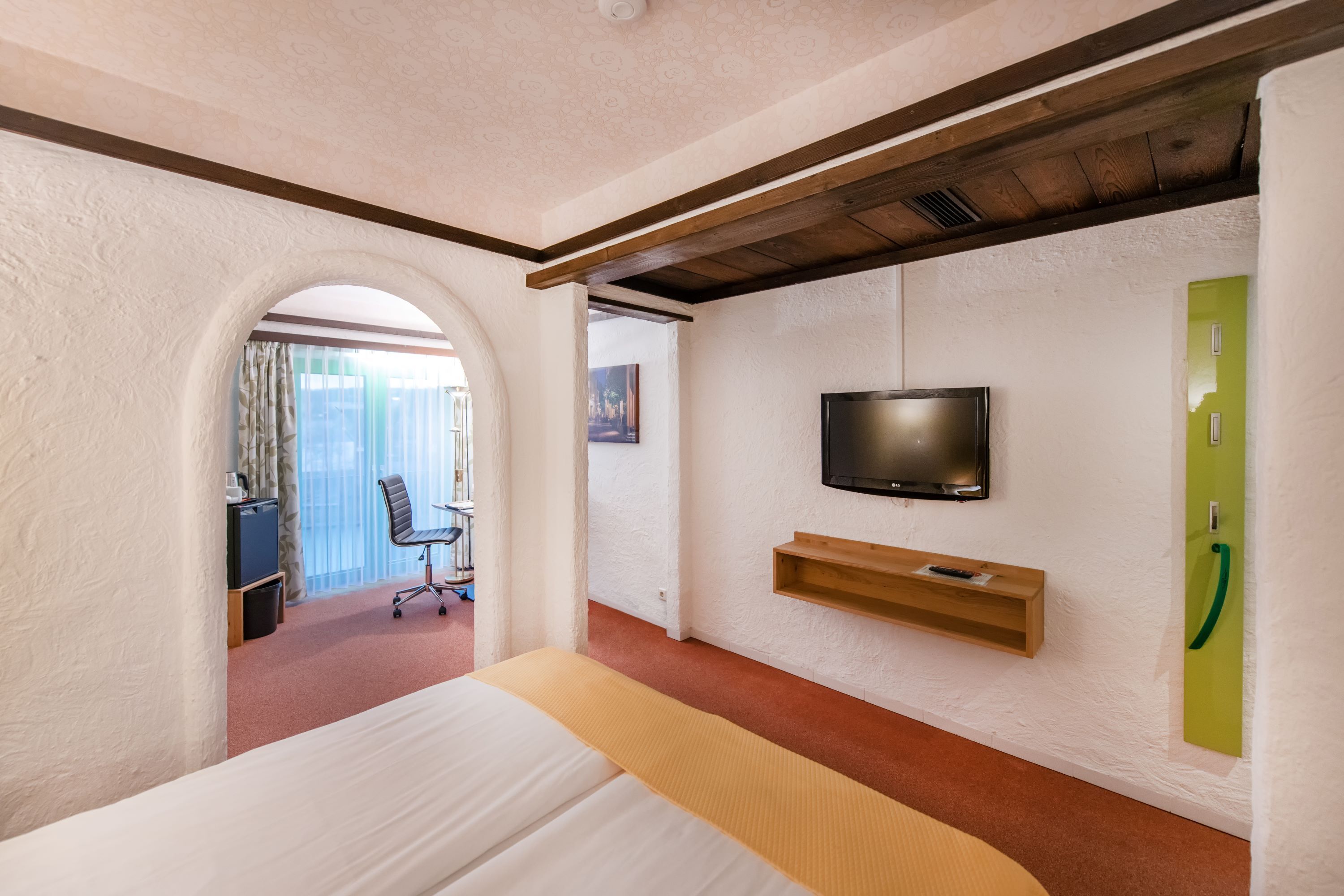 Habitación de hotel con cama, televisión montada en la pared, estante de madera y un elemento decorativo en la pared verde.