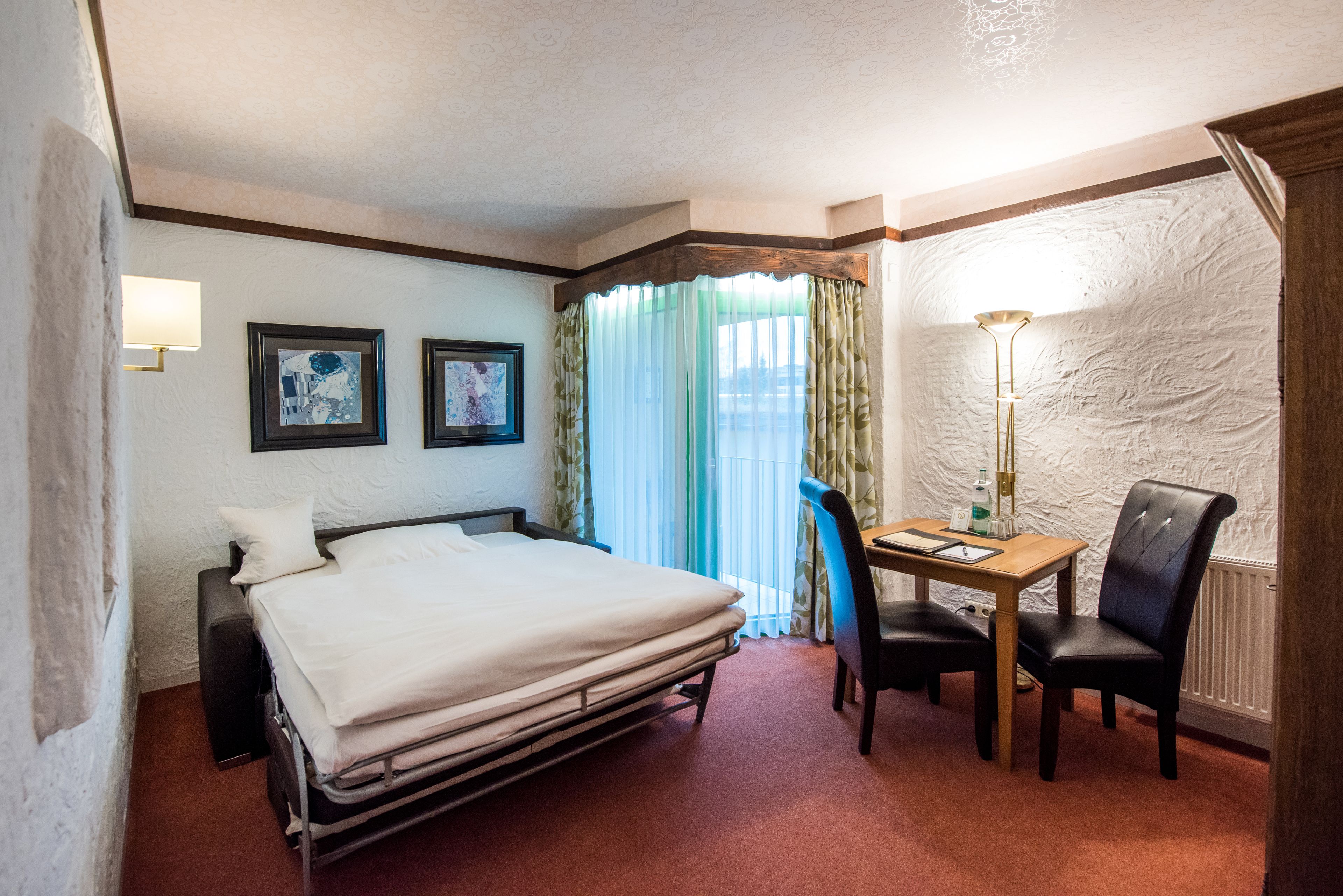 Ein Hotelzimmer mit einem Bett, einem Schreibtisch mit Stuhl, einer Lampe und zwei gerahmten Bildern an der Wand.