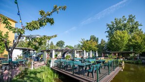 Un restaurante a la orilla del río con terraza al aire libre sobre una cubierta de madera, rodeado de follaje y un cielo azul despejado.