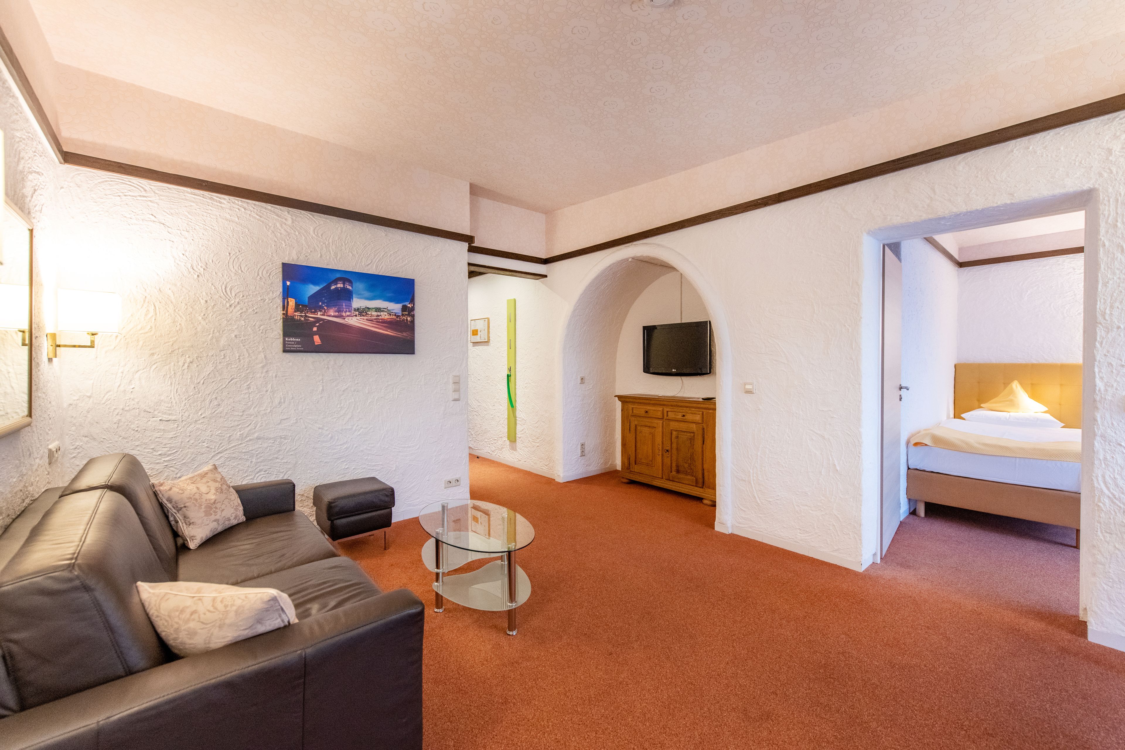 A modern hotel room with a sofa, a glass coffee table, a TV, and a bed.