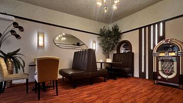 A room with a brown carpet, a brown sofa, a brown armchair, a small table, a potted plant, a mirror, a wall-mounted radio, and a chandelier.