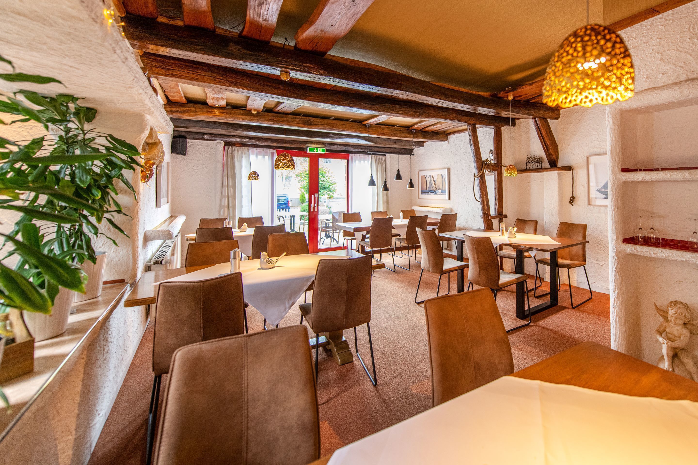Un restaurante con vigas de madera, paredes blancas y una puerta roja. Mesas y sillas dispuestas para comer.