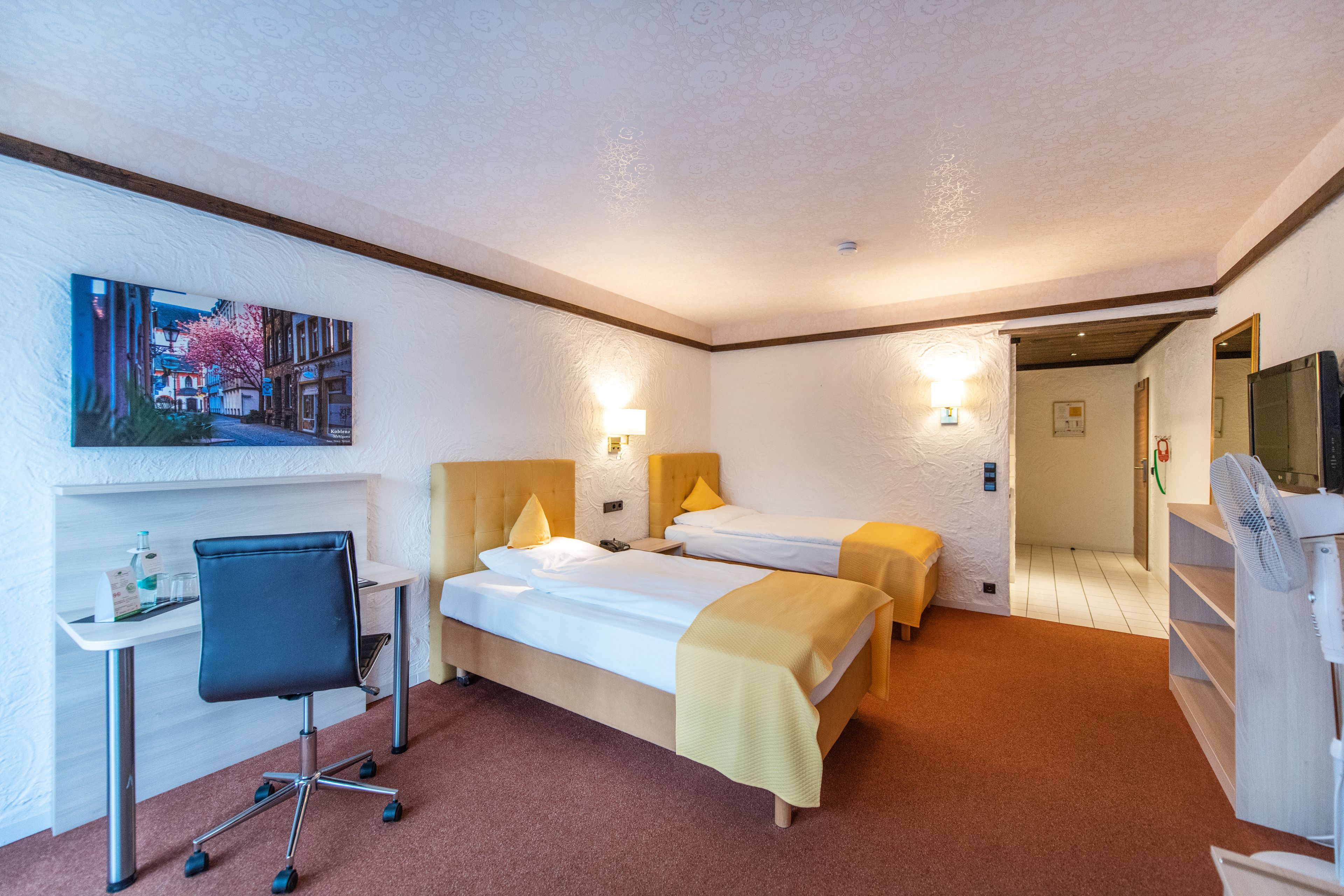 A hotel room with two beds, a desk with a chair, a painting on the wall, and a TV on the right.