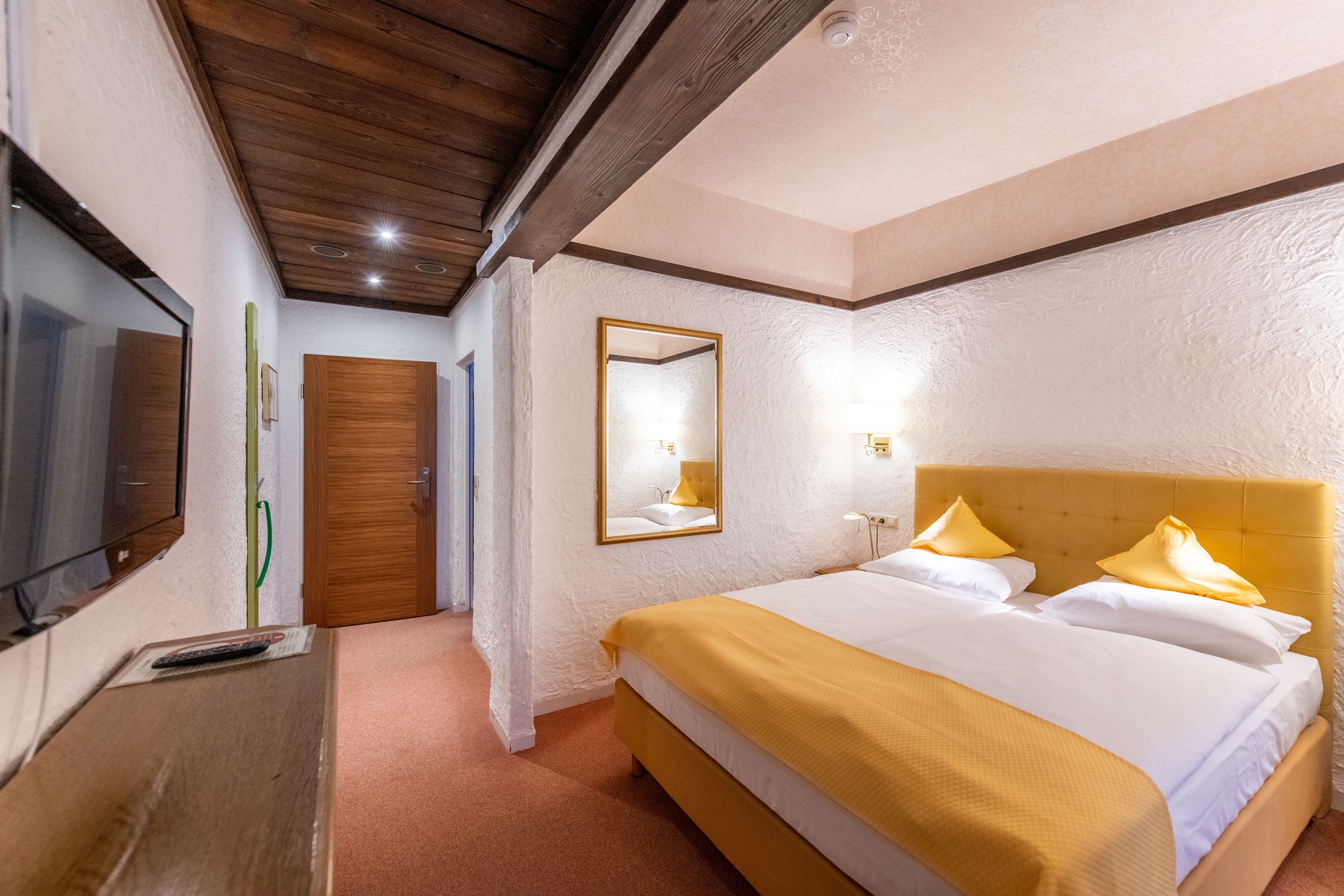 Una habitación de hotel con techo de madera, una cama grande con ropa de cama blanca y amarilla, un televisor y un espejo en la pared.